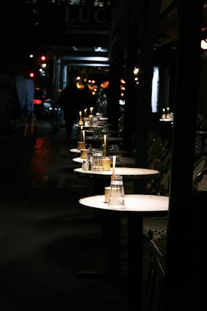 A dimly lit outdoor dining area on a city street, with several small round tables lined up. Each table has an unlit candle and upside-down glasses, suggesting a restaurant setup. The background shows blurred pedestrians and softly glowing city lights, creating a serene nighttime atmosphere.