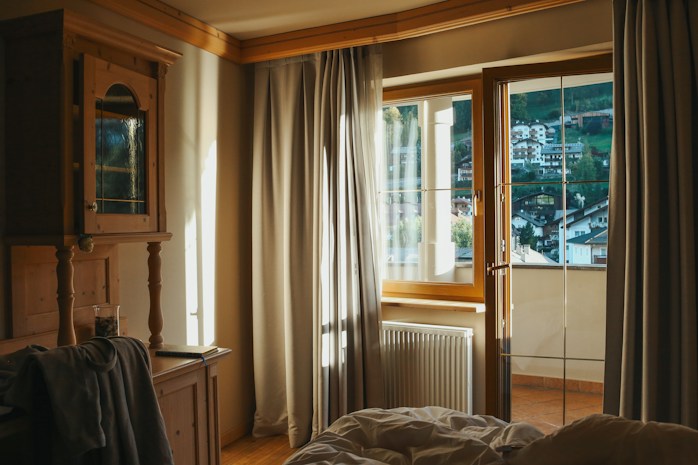 Warm sunlight filters through large windows with sheer curtains, illuminating a cozy room that features wooden furniture and soft bedding. Beyond the glass, a picturesque view of a hillside town with clustered houses is visible, framed by lush green trees.