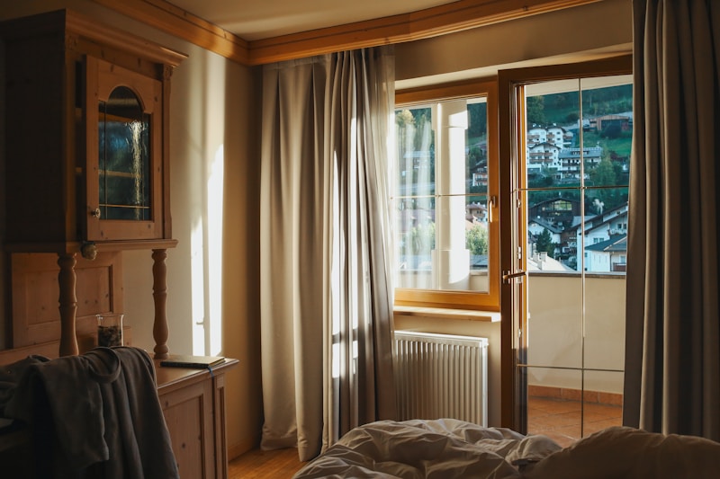 Warm sunlight filters through large windows with sheer curtains, illuminating a cozy room that features wooden furniture and soft bedding. Beyond the glass, a picturesque view of a hillside town with clustered houses is visible, framed by lush green trees.