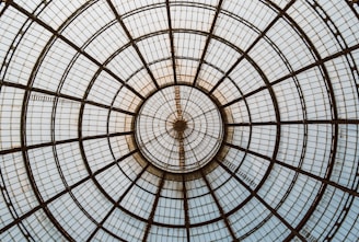 A symmetrical dome structure with a radial pattern composed of metal and glass panels converging at a central point. The design features concentric circles and straight lines that create a geometric and architectural appearance.