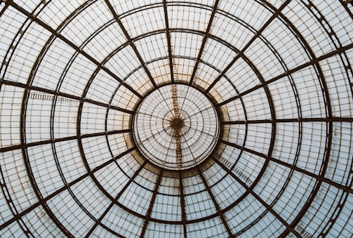 A symmetrical dome structure with a radial pattern composed of metal and glass panels converging at a central point. The design features concentric circles and straight lines that create a geometric and architectural appearance.
