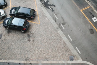 Aerial view of a parking area with three parked cars. The cars are dark-colored and parked on a cobblestone surface, marked with yellow lines. A gray road runs adjacent to the parking area, with visible road markings and a pedestrian crossing symbol.