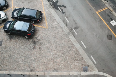 Aerial view of a parking area with three parked cars. The cars are dark-colored and parked on a cobblestone surface, marked with yellow lines. A gray road runs adjacent to the parking area, with visible road markings and a pedestrian crossing symbol.
