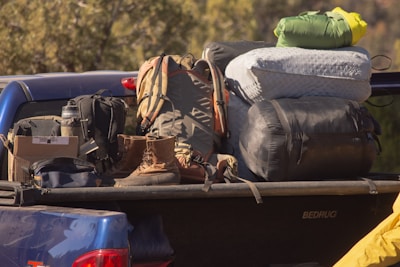 A collection of camping gear is loaded onto the back of a blue pickup truck. Among the items visible are backpacks, a pair of hiking boots, a water bottle, a cardboard box, and several sleeping bags. The gear is assorted in an organized manner, hinting at preparation for a camping trip.