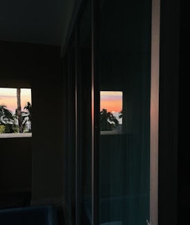 The image captures a tranquil sunset view seen through a window, with palm trees silhouetted against the vibrant orange and pink sky. The reflection on the glass adds a symmetrical element, enhancing the serene atmosphere.