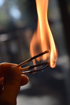 A close-up of two burning matches held together with a warm, bright flame rising from the match heads. The background is blurred, enhancing the focus on the flame and the hand holding the matches.