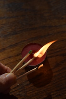 A hand is holding a group of three matchsticks, one of which is ignited. The flame is bright against the dark, textured wooden surface. Behind the flame, there is a red tealight candle in a holder with a small amount of wax, casting a shadow on the surface.