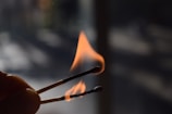 A close-up of two burning matchsticks against a dark, blurry background. The flames are prominent, with one matchstick visibly flaming while the other shows signs of burning out.