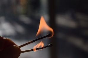 A close-up of two burning matchsticks against a dark, blurry background. The flames are prominent, with one matchstick visibly flaming while the other shows signs of burning out.