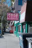 A street scene featuring a red sign with both Chinese characters and the text 'Gourmet Food Inc.' prominently displayed. The sign is attached to a green building, which has several windows. A tree lines the sidewalk along with a garbage bin and a commercial establishment. In the background, other buildings and a street lined with trees are visible.