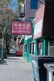 A street scene featuring a red sign with both Chinese characters and the text 'Gourmet Food Inc.' prominently displayed. The sign is attached to a green building, which has several windows. A tree lines the sidewalk along with a garbage bin and a commercial establishment. In the background, other buildings and a street lined with trees are visible.