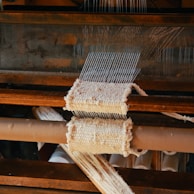 A close-up view of a wooden loom in the process of weaving fabric. The loom has multiple parallel threads and partially woven material is visible. The texture of the textile and the wooden components of the equipment are prominent.