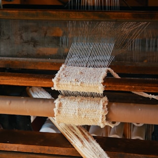 A close-up view of a wooden loom in the process of weaving fabric. The loom has multiple parallel threads and partially woven material is visible. The texture of the textile and the wooden components of the equipment are prominent.