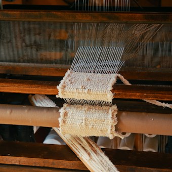 A close-up view of a wooden loom in the process of weaving fabric. The loom has multiple parallel threads and partially woven material is visible. The texture of the textile and the wooden components of the equipment are prominent.