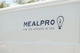 The side of a white vehicle displays the meal service logo 'MEALPRO' with the tagline 'For the Athlete in You'. Next to the text is a graphic combining a lightbulb and a fork.