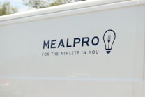 The side of a white vehicle displays the meal service logo 'MEALPRO' with the tagline 'For the Athlete in You'. Next to the text is a graphic combining a lightbulb and a fork.