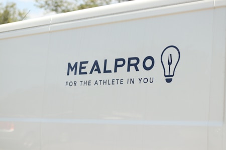 The side of a white vehicle displays the meal service logo 'MEALPRO' with the tagline 'For the Athlete in You'. Next to the text is a graphic combining a lightbulb and a fork.