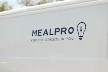 The side of a white vehicle displays the meal service logo 'MEALPRO' with the tagline 'For the Athlete in You'. Next to the text is a graphic combining a lightbulb and a fork.