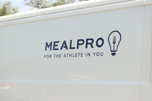 The side of a white vehicle displays the meal service logo 'MEALPRO' with the tagline 'For the Athlete in You'. Next to the text is a graphic combining a lightbulb and a fork.