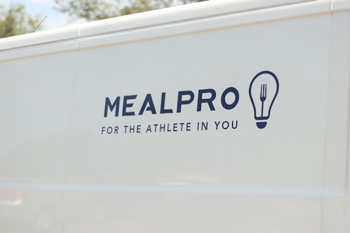 The side of a white vehicle displays the meal service logo 'MEALPRO' with the tagline 'For the Athlete in You'. Next to the text is a graphic combining a lightbulb and a fork.