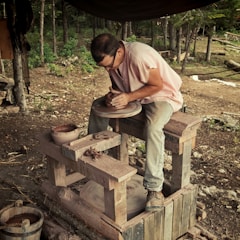 A person focused on shaping clay on a potter's wheel in an outdoor setting surrounded by trees and natural materials. The individual's hands are actively molding the clay, and various pottery tools and materials are scattered around.