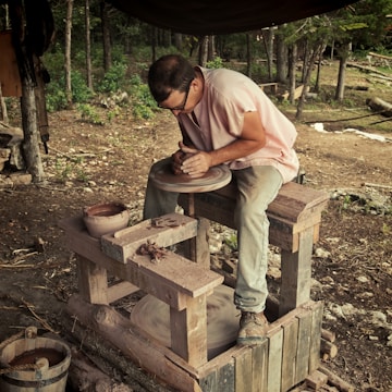 A person focused on shaping clay on a potter's wheel in an outdoor setting surrounded by trees and natural materials. The individual's hands are actively molding the clay, and various pottery tools and materials are scattered around.