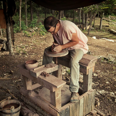 An artisan shaping clay on a potter’s wheel in a sunlit studio filled with plants.