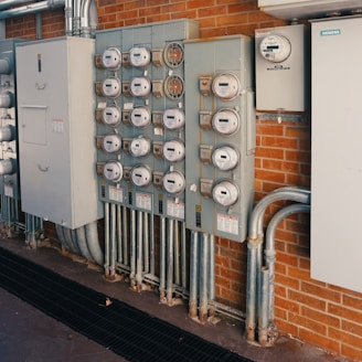A row of multiple electrical meters and panels is mounted on a brick wall. The setup includes several round meters with clear covers, metal conduits, and electrical boxes. The equipment is organized and securely installed.