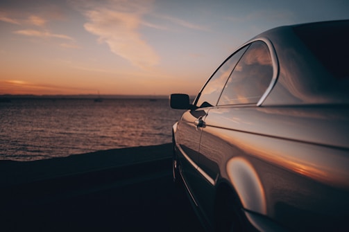 A sleek luxury car parked by the coast at sunset, reflecting the warm colors of the sky