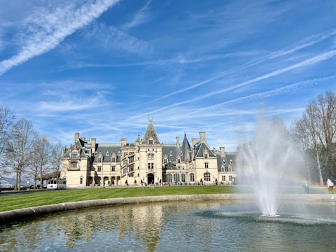 A grand, historic mansion with a steeply pitched roof and multiple chimneys against a clear blue sky. A large fountain is prominently featured in the foreground, surrounded by a circular water basin. Leafless trees frame the scene, adding a touch of natural elegance.