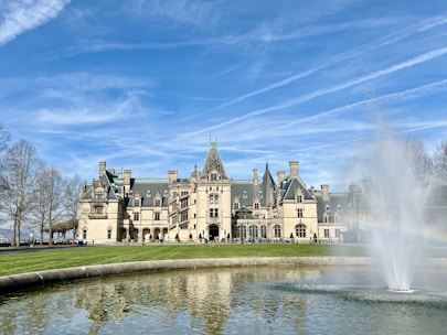 A large, grandiose mansion sits under a clear blue sky with streaks of white clouds. In the foreground, a round fountain with a gentle spray of water reflects the building and surrounding area. The architecture of the mansion is elaborate, with numerous windows, chimneys, and a central tower. Bare trees frame the image, indicating an early spring setting.