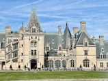 A large and ornate stone mansion with Gothic architectural elements, including tall towers, pointed spires, and detailed window and door decorations. The building has several chimneys and a glass atrium in the center. People are walking and standing in front of the mansion, with a green lawn in the foreground and a clear blue sky above.