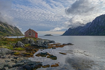 A solitary red wooden cabin sits on a rocky shoreline, surrounded by rugged mountains and a calm body of water. The sky is overcast with scattered clouds, creating a serene and tranquil atmosphere. Vegetation and rocks are visible along the coast, adding to the rustic feel of the scene.