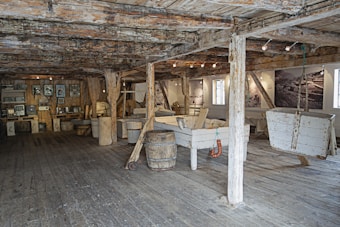 A rustic interior with wooden beams and floorboards, featuring vintage machinery and equipment along one wall. Large wooden barrels and troughs are scattered throughout the space, contributing to an industrial, historical atmosphere. The walls display black-and-white photographs and the room is lit with spotlights.