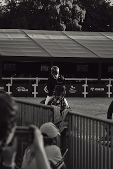 A black and white photograph featuring a person wearing riding gear, seated on a horse. The scene appears to be at an equestrian event, with a backdrop of a large tent and sponsor logos. In the foreground, blurred figures take photos, focusing on the rider and horse. Trees are visible in the background, adding depth and context to the setting.