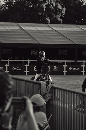 A black and white photograph featuring a person wearing riding gear, seated on a horse. The scene appears to be at an equestrian event, with a backdrop of a large tent and sponsor logos. In the foreground, blurred figures take photos, focusing on the rider and horse. Trees are visible in the background, adding depth and context to the setting.