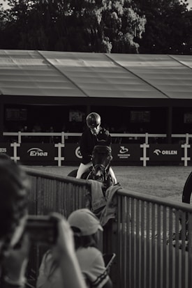 A black and white photograph featuring a person wearing riding gear, seated on a horse. The scene appears to be at an equestrian event, with a backdrop of a large tent and sponsor logos. In the foreground, blurred figures take photos, focusing on the rider and horse. Trees are visible in the background, adding depth and context to the setting.