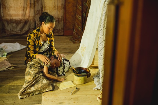 A woman in a patterned dress sits on a wooden floor, holding a young child who is resting on her lap. The setting appears to be indoors with a rustic, warm ambiance. There are textiles with intricate designs around them, as well as a bowl and a piece of paper nearby.