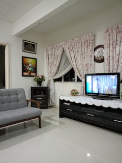 A cozy living room with a light gray fabric sofa, a black wooden coffee table with a floral arrangement, and a wall-mounted TV displaying a scene. The walls feature decorative art pieces, and floral-patterned curtains hang over a window. A clock is positioned above the TV.