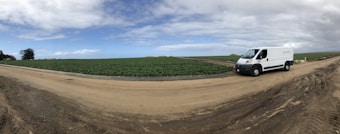 A large field with rows of green crops stretches under a partly cloudy sky. A white van is parked on a dirt path that intersects the field, with some trees visible in the distance.
