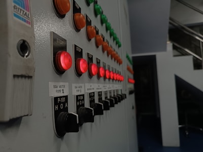 A series of control panel buttons are displayed in rows, featuring illuminated red, orange, and green push buttons. Labels next to the buttons indicate functions such as 'STOP' and 'RAW WATER PUMP.' The perspective shows the panel extending into the distance, suggesting an industrial or mechanical setting with metal surroundings.