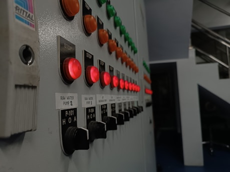 A series of control panel buttons are displayed in rows, featuring illuminated red, orange, and green push buttons. Labels next to the buttons indicate functions such as 'STOP' and 'RAW WATER PUMP.' The perspective shows the panel extending into the distance, suggesting an industrial or mechanical setting with metal surroundings.