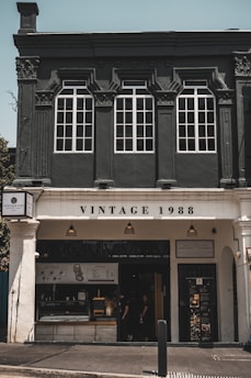 The image features a vintage-style cafe with a dark fa&ccedil;ade and classic architectural details, including large white-framed windows. The sign reads 'Vintage 1988' above the entrance, flanked by three warm-hued exterior lights. The open front reveals a menu board and a glimpse of the interior with chairs and people inside.