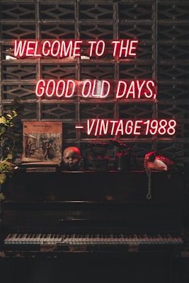 A dark, vintage setting features a neon sign with red letters stating 'Welcome to the good old days, - Vintage 1988' against a patterned brick wall. Below the sign is an antique upright piano with various items on top, including a doll's head, a vintage telephone, and framed art. Some foliage adds a touch of nature to the scene.