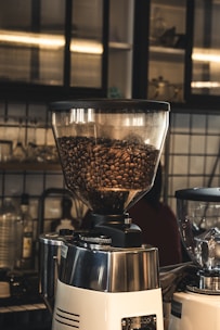 A coffee grinder filled with brown coffee beans is positioned in a cozy cafe setting, surrounded by a variety of glass bottles and utensils on shelves. The background features tiled walls and soft ambient lighting, adding to the warm and inviting atmosphere.