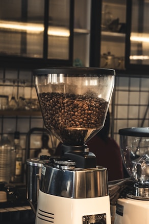 A coffee grinder filled with brown coffee beans is positioned in a cozy cafe setting, surrounded by a variety of glass bottles and utensils on shelves. The background features tiled walls and soft ambient lighting, adding to the warm and inviting atmosphere.