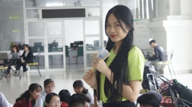A young woman with long dark hair, wearing a lime green top and holding some papers, stands smiling in a bright indoor setting. Several other people are seated around in what appears to be a casual, social environment with white walls and glass doors.