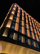 An illuminated building at night with a unique architectural design, featuring vertical orange and yellow panels and numerous windows. The lighting accentuates the textured patterns on the facade, creating a striking contrast against the dark sky.
