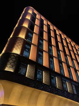 An illuminated building at night with a unique architectural design, featuring vertical orange and yellow panels and numerous windows. The lighting accentuates the textured patterns on the facade, creating a striking contrast against the dark sky.
