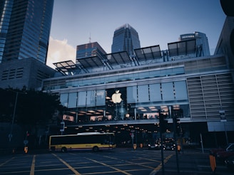 A modern cityscape featuring a prominent commercial building with a large Apple logo on the facade. The structure is composed of glass and metal, with a spacious second-floor window showcasing the interior lighting. A yellow bus is seen crossing the street, surrounded by traffic lights and road markings. Skyscrapers tower in the background, adding to the urban atmosphere.